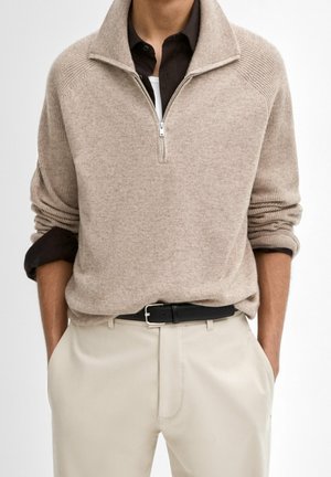 Man wearing beige half-zip sweater over black shirt with sleeves rolled up, tucked into light beige pants with black belt. Hands in pockets.