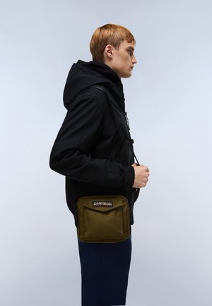 Geantă de umăr verde măsliniu cu clapetă, având o insignă cu logo. Poartă peste o jachetă neagră, afișată pe un fundal deschis.