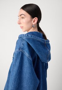Överdimensionerad blå denimhuva med dragsko, synlig sömnad och mjuk textur. Modellen visas från sidan, örhängen synliga.