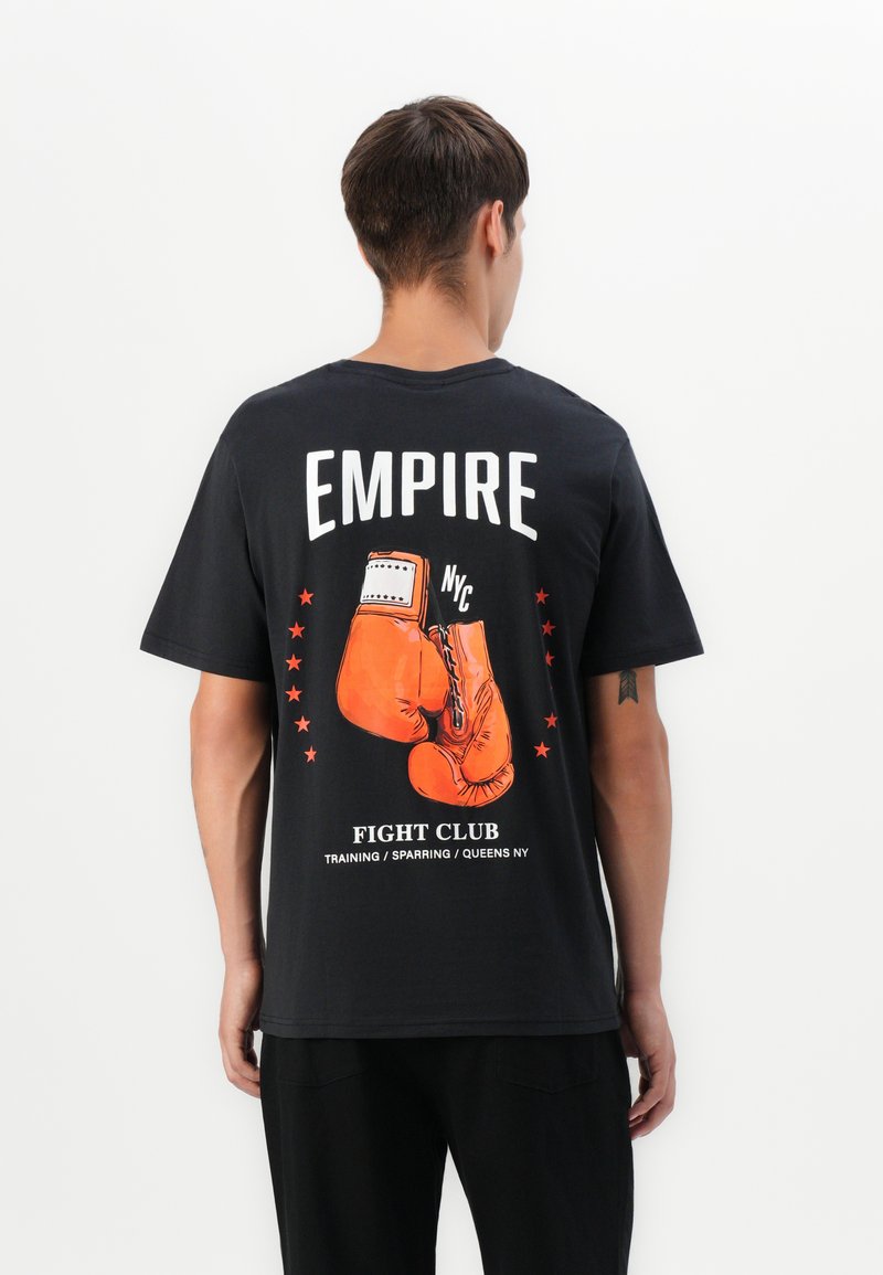 Ung mand iført sort t-shirt med orange boksehandsker grafik og teksten "Empire Fight Club, Træning/Sparring/Queens NY" på ryggen.