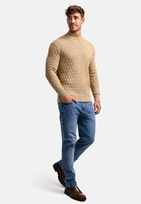 Pull en tricot beige avec un motif texturé, col haut, associé à un jean bleu clair et des bottines marron à bout rond.