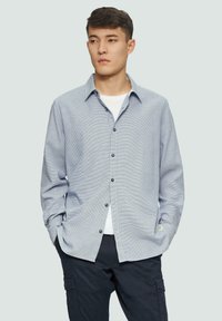 Chemise à manches longues avec un motif texturé bleu et blanc, col classique ; associée à un pantalon sombre.