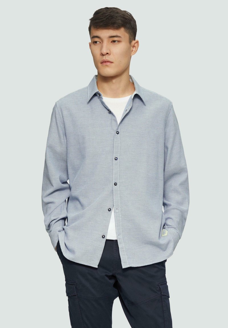 Chemise à manches longues avec un motif texturé bleu et blanc, col classique ; associée à un pantalon sombre.