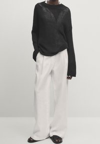 Pull en maille noire à coupe ample avec manches larges, associé à un pantalon blanc à jambes larges en tissu léger. Sandales noires.