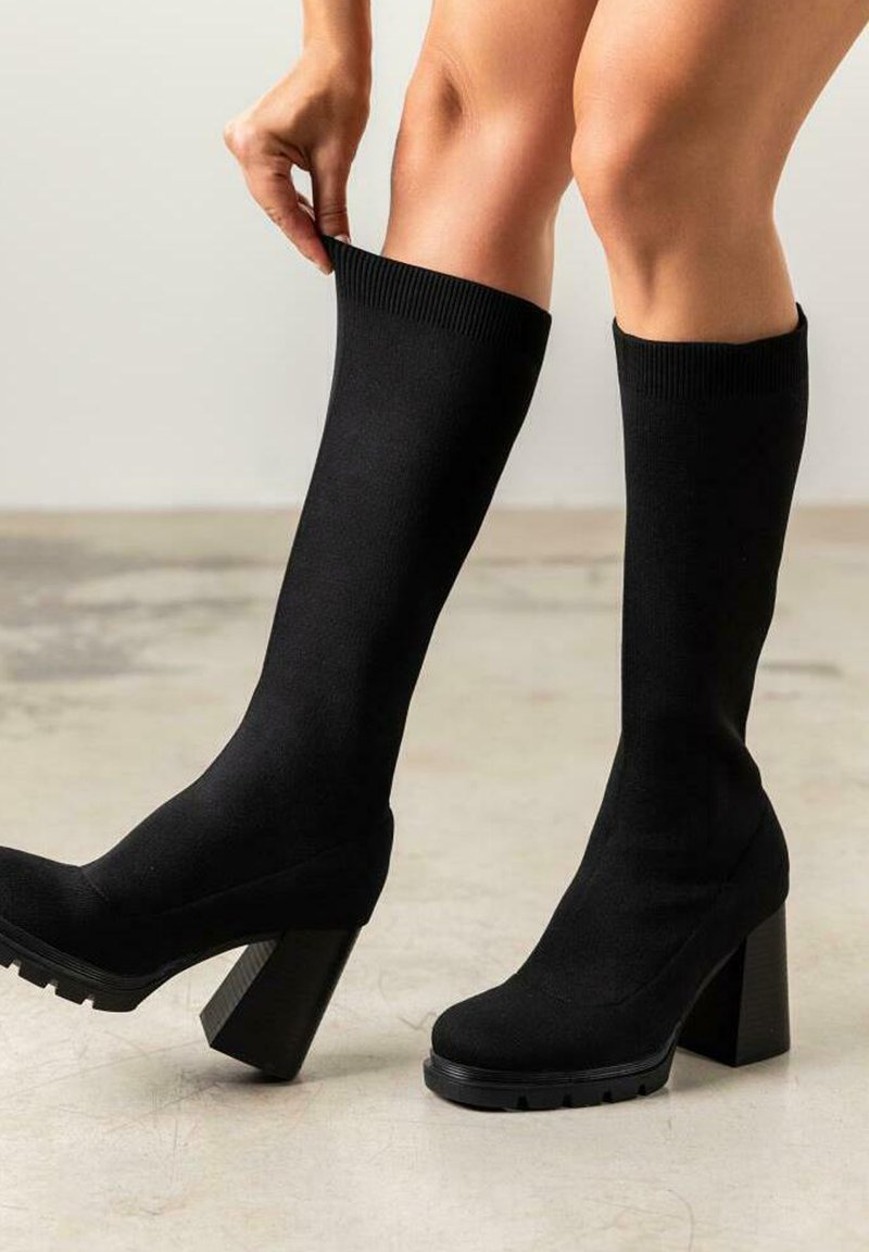 Botas negras hasta la rodilla con un upper de tela elástica ajustada, tacón grueso bloque y suela texturizada para agarre. Sin patrones o acentos visibles.