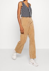 Blusa senza maniche a righe blu navy e bianche con bottoni, abbinata a pantaloni a gamba larga in velluto a coste beige a vita alta e sneakers bianche.