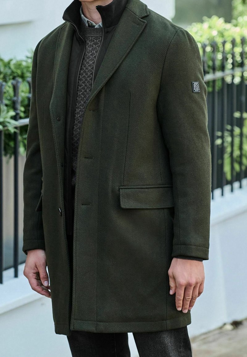 Homme portant un pardessus vert foncé sur une veste zippée noire et un pull à motifs, debout à l'extérieur près d'une clôture noire et de verdure.