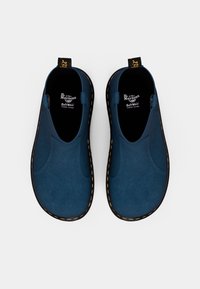 Blue suede slip-on shoes with yellow stitching and black rubber soles. SoftWear technology label visible inside. Smooth, rounded design.