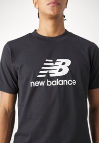 Czarny t-shirt z bawełny z okrągłym dekoltem, z dużym białym logotypem „New Balance” i graficznym wzorem w paski. Gładka tekstura, krótkie rękawy.