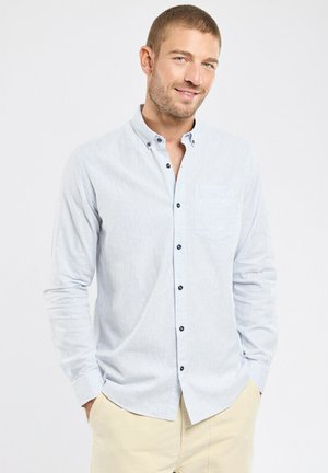 Homme les mains dans les poches, vêtu d'une chemise bleu clair à boutons et d'un pantalon beige, souriant légèrement devant un fond blanc uni.