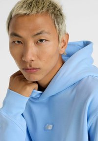 Lyseblå hoodie med en blød tekstur. Har et logo på forsiden og fastgørelse til hætten. Synlige ribbede manchetter. Model vist med hånden tæt på ansigtet.