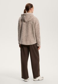 Giacca beige pelosa con cappuccio, maniche larghe e orlo arrotondato, abbinata a pantaloni larghi marroni e sneakers bianche. Dettaglio del tessuto testurizzato visibile.