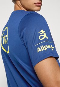 Blå atletisk t-shirt lavet af glat stof, med et gult logo og tekst på ærmet. Korte ærmer med en tætsiddende design.