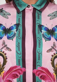 Roze shirt met teal en donkerpaarse verticale strepen, versierd met blauwe vlinders, libellen, roze veren en de tekst "Versace Jeans Couture".