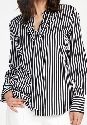 Kobieta ubrana w czarno-białą, pionowo paskowaną bluzkę z długim rękawem z wiązaniem z przodu, w połączeniu z białymi spodniami, ręka na biodrze.