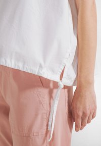 Top blanco de algodón con lazos laterales y un dobladillo ligeramente curvado, llevado sobre pantalones rosa claro con un bolsillo lateral y tejido texturizado.