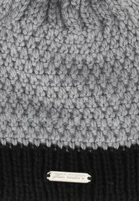 Stickad mössa med en texturerad ljusgrå överdel och en svart ribbad kant, med en silverfärgad logotag sydd på den svarta delen.