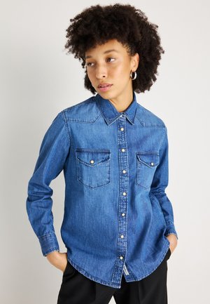 Camicia in denim blu con colletto, due tasche sul petto e chiusura con bottoni. Presenta maniche lunghe e un orlo curvo. Tessuto testurizzato.