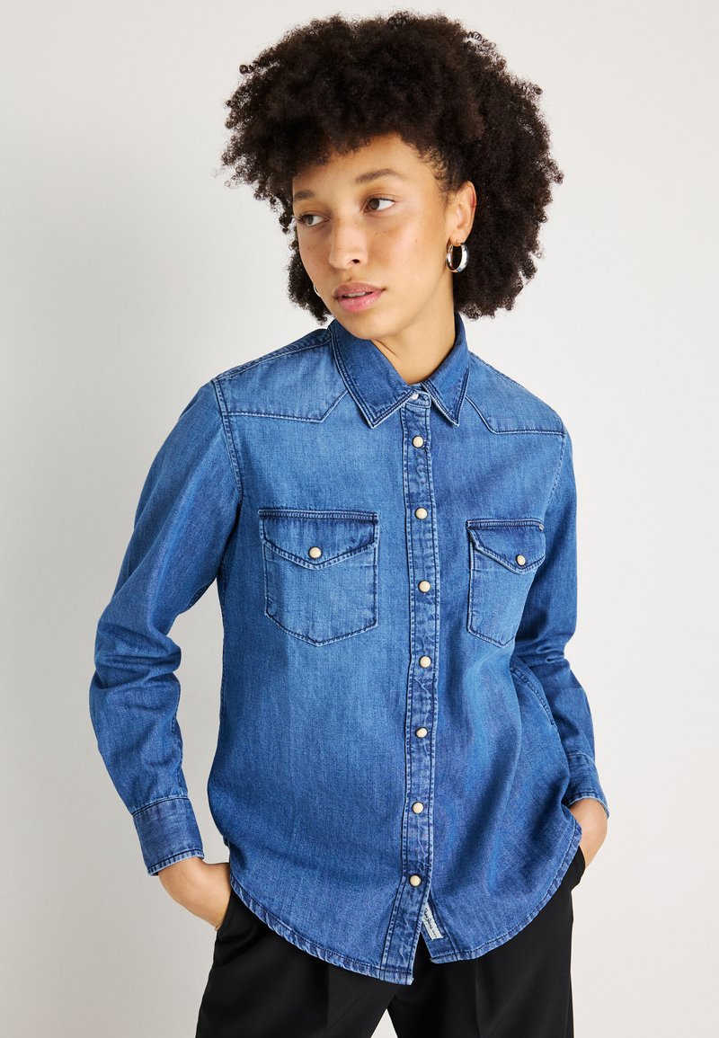 Camisa de ganga azul com colarinho, dois bolsos no peito e fechos de botão. Tem mangas longas e uma bainha curva. Tecido texturizado.