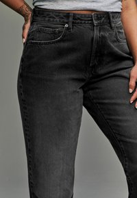 Jeans de mezclilla negra con alto cintura, diseño de cinco bolsillos y costuras visibles. Textura suave, acabados ligeramente desgastados y botón de metal.