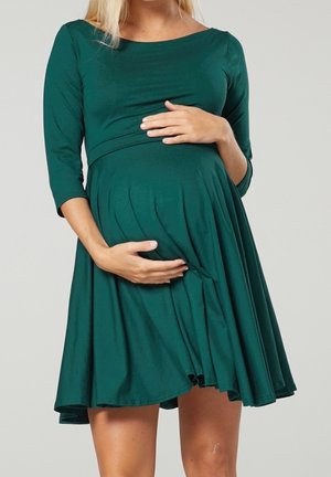 Schwangere Frau in einem grünen, knielangen Kleid mit dreiviertel Ärmeln, die sanft ihren Bauch vor einem schlichten Hintergrund hält.