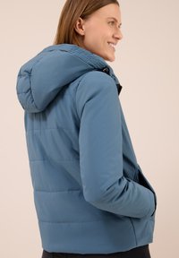 Giacca a vento azzurra chiara con cappuccio, dotata di pannelli cuciti, tasche laterali e chiusura con zip. Tessuto liscio e resistente.