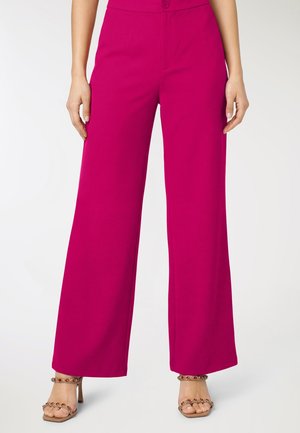 Donna che indossa pantaloni magenta a vita alta e gamba larga abbinati a sandali con tacco aperto decorati con dettagli rossi e dorati.