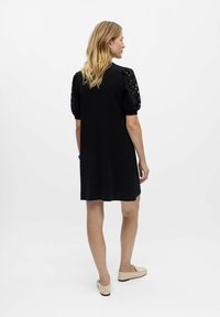 Schwarzes Kurzarmkleid mit strukturierten Puffärmeln und Ausschnitten, knielang, lockere Passform, getragen mit hellen Schuhen.