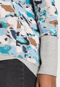 Gros plan sur une personne portant un pull à motifs abstraits multicolores avec des poignets et un ourlet gris côtelés, associé à un pantalon sombre.