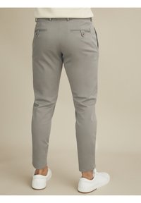 Mies, joka on pukeutunut vaaleanharmaisiin slim-fit-housuihin ja valkoisiin lenkkareihin, seisoo yksinkertaista beigeä taustaa vasten, nähtynä takaa.