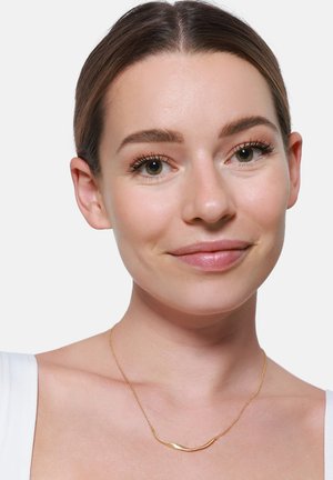 Collana d'oro con pendente a barra curva, catena delicata e finitura lucida, indossata su una clavicola nuda su uno sfondo chiaro.