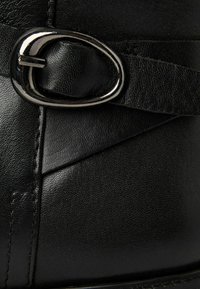 Stivale alla caviglia in pelle nera con punta arrotondata, fascia decorativa con fibbia ovale in argento e superficie texturizzata con cuciture visibili.