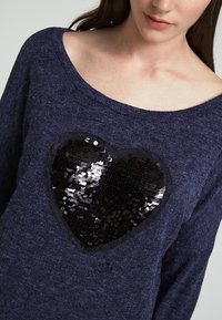 Marinblå stickad tröja med vid halsringning, med ett stort svart paljetthjärta i mitten, strukturerad tyg och mjuk känsla.
