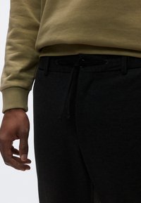 Μαύρο παντελόνι φόρμας με κορδόνι και ελαστική μέση, συνδυασμένο με φούτερ σε ελαιόχρωμο πράσινο. Κοντινή εικόνα της υφής του υφάσματος και της ρ αφής.