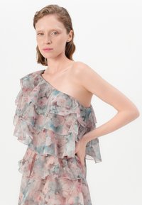 Kvinna med kort vågigt hår som bär en axelbandslös, lager-på-lager blommig klänning med volanger, poserar med en hand på höften framför en vit bakgrund.