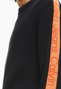 Schwarzer Strickpullover mit einem schmalen, orangen Streifen und dem Schriftzug "Calvin Jeans" entlang des Ärmels. Geripptes Material und Rundhalsausschnitt.