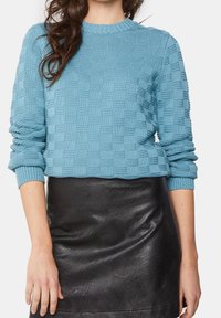 Pull en maille texturée bleu clair avec col rond et poignets côtelés, associé à une jupe en simili cuir noir, lisse et ajustée.