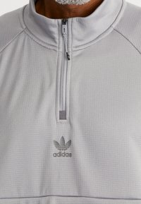 Světlé šedé pulovr s polovičním zipem, texturovanou tkaninou a jemným logem adidas na hrudi. Má stojá ček a šicí detaily.
