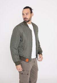 Olijfgroene bomberjack met ribgebreide manchetten en zoom, voorzien van een rits en oranje labeldetail aan de voorkant, gedragen over een wit shirt.