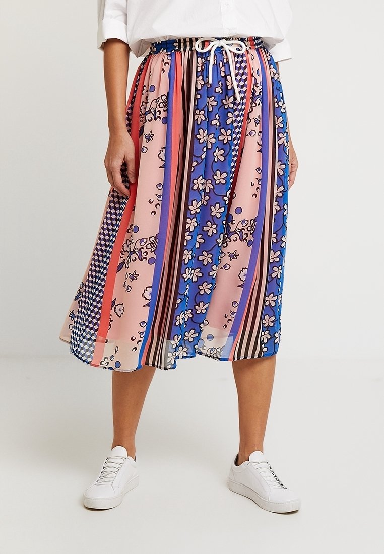 Femme portant une jupe midi colorée à motifs floraux et rayures, taille blanche avec cordon, associée à des baskets blanches et un haut blanc.