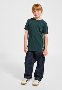 Grøn kortærmet t-shirt med logoaccent, kombineret med løstsiddende mørk denimjeans og hvide sneakers med lysebrune såler.