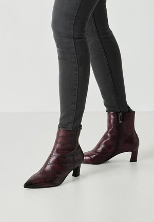 Vrouw die donkergrijze skinny jeans draagt en bordeauxrode enkellaarsjes met een puntige neus, een lage blokhak en een krokodillenpatroon.