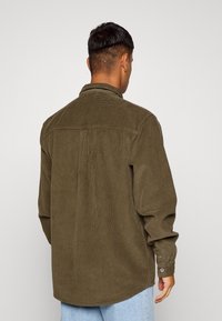 Olivově zelená košile z manšestru, s dlouhými rukávy, knoflíkovými manžetami a klasickým límcem. Má vertikální žebrovanou strukturu a zakřivený spodní lem.