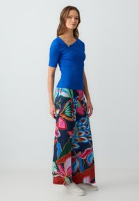 Blauw aansluitend topje met korte mouwen, gecombineerd met wijde broek met een levendige bloemenprint in roze, groen en blauw. Witte sneakers.