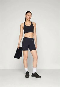 Un crop top et un short de sport noirs, associés à des baskets noires, des chaussettes blanches et une serviette noire drapée sur un bras. Matériel lisse et ajusté.