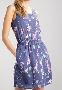 Fioletowa sukienka w kwiaty o bezrękawnikowym kroju, elastycznym pasie, małych kieszeniach, różowych kwiatach i zielonych liściach na miękkim tle.