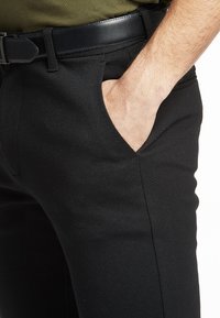 Zwarte broek van gladde stof, voorzien van zijzakken, een vlakke voorkant en een slanke pasvorm. Draagt een zwarte riem.