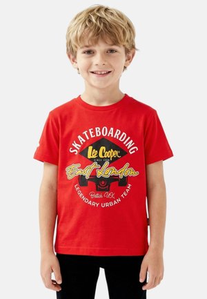 Garçon blond souriant, portant un t-shirt rouge avec des textes et des graphismes blancs et jaunes sur le thème du skateboard, debout devant un fond clair uni.