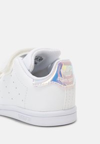 Sneaker in pelle bianca con lati perforati e linguetta olografica sul tallone con la dicitura "stan smith". Suola piatta e dettaglio con strap in velcro.