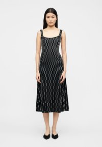 Femme portant une robe midi noire sans manches avec un décolleté carré et des lignes blanches pointillées verticales, debout devant un fond uni.